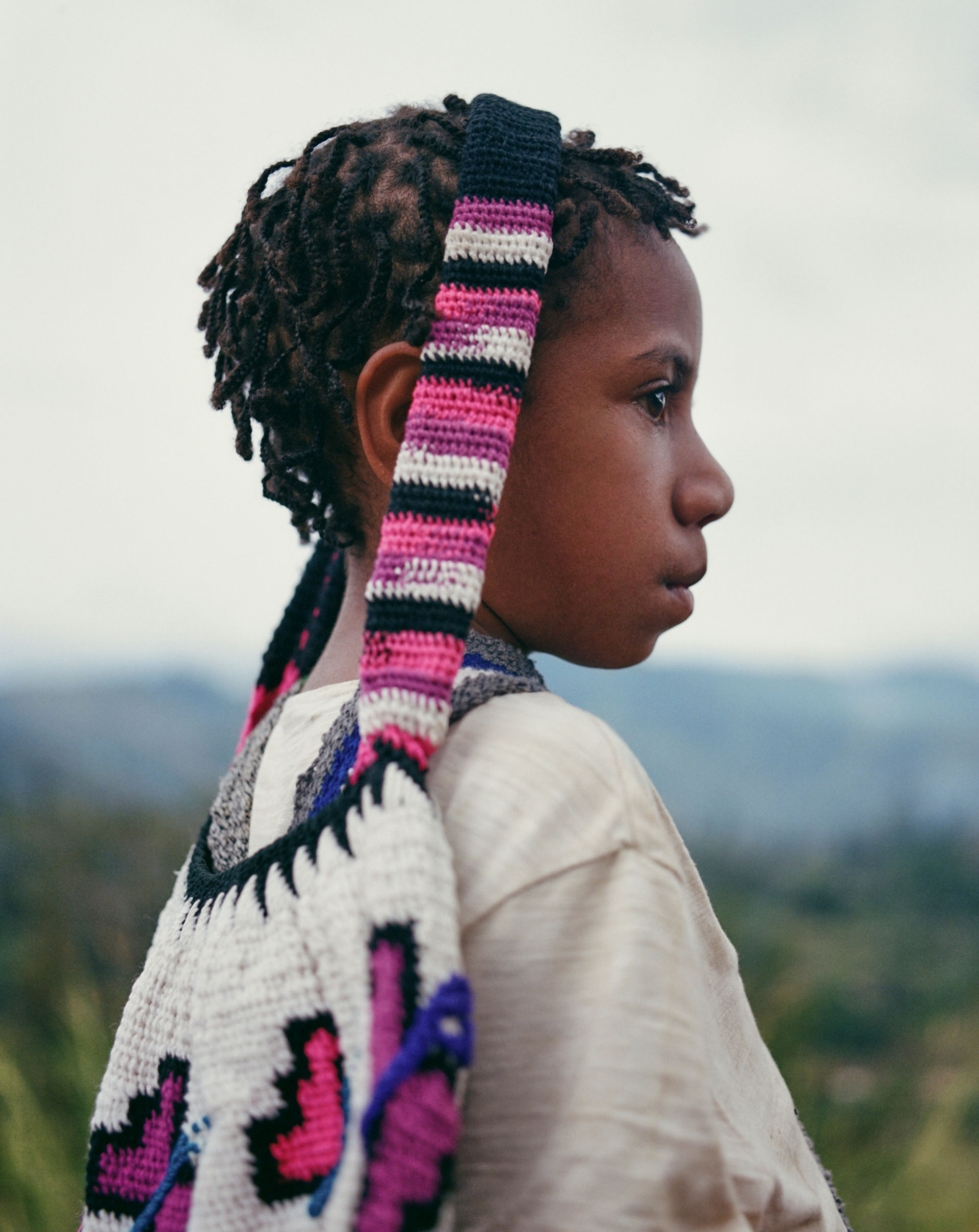 Profile shot of a contemplative child with dreadlocks wearing a light shirt and carrying a colorful crochet bag in a natural setting.