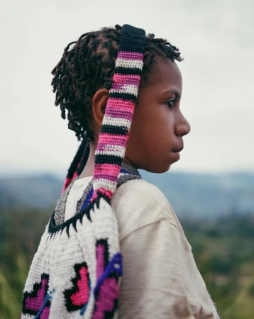 Profile shot of a contemplative child with dreadlocks wearing a light shirt and carrying a colorful crochet bag in a natural setting.