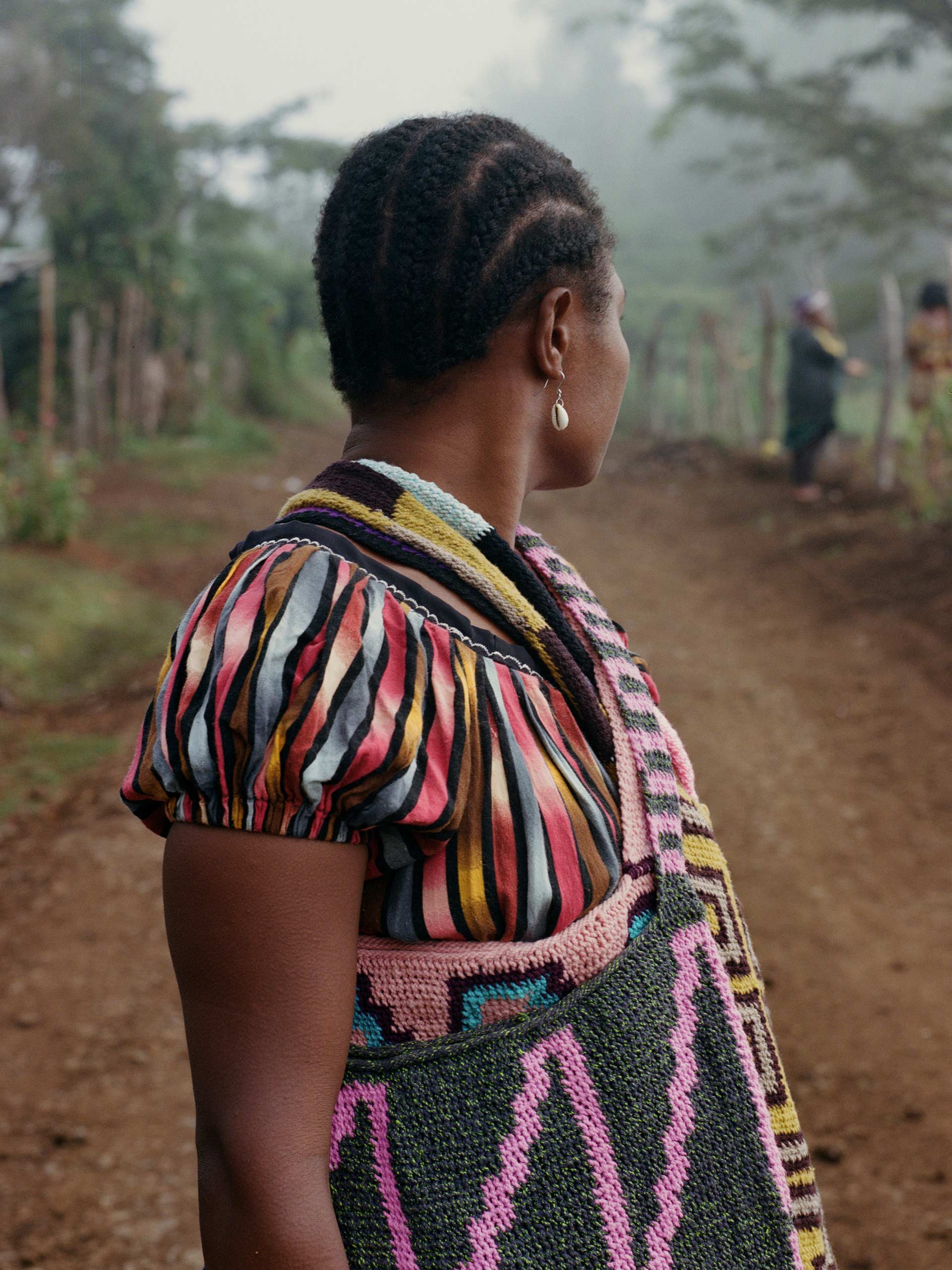 Woman stands on dirt path in rural area, seen from the side, wearing a colorful blouse, shoulder bag, and shell earrings in a medium shot.