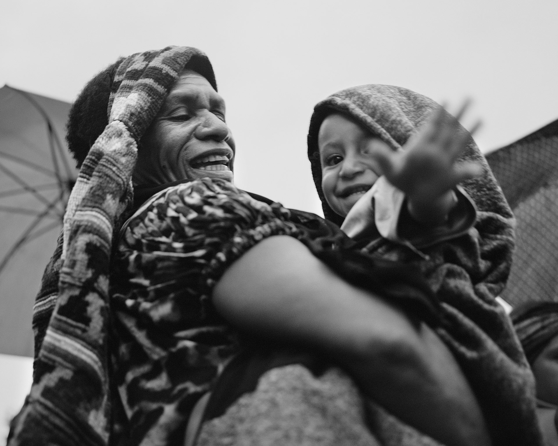 Black and white shot of a smiling woman holding a young child wrapped in a patterned hooded blanket, both in an outdoor setting.