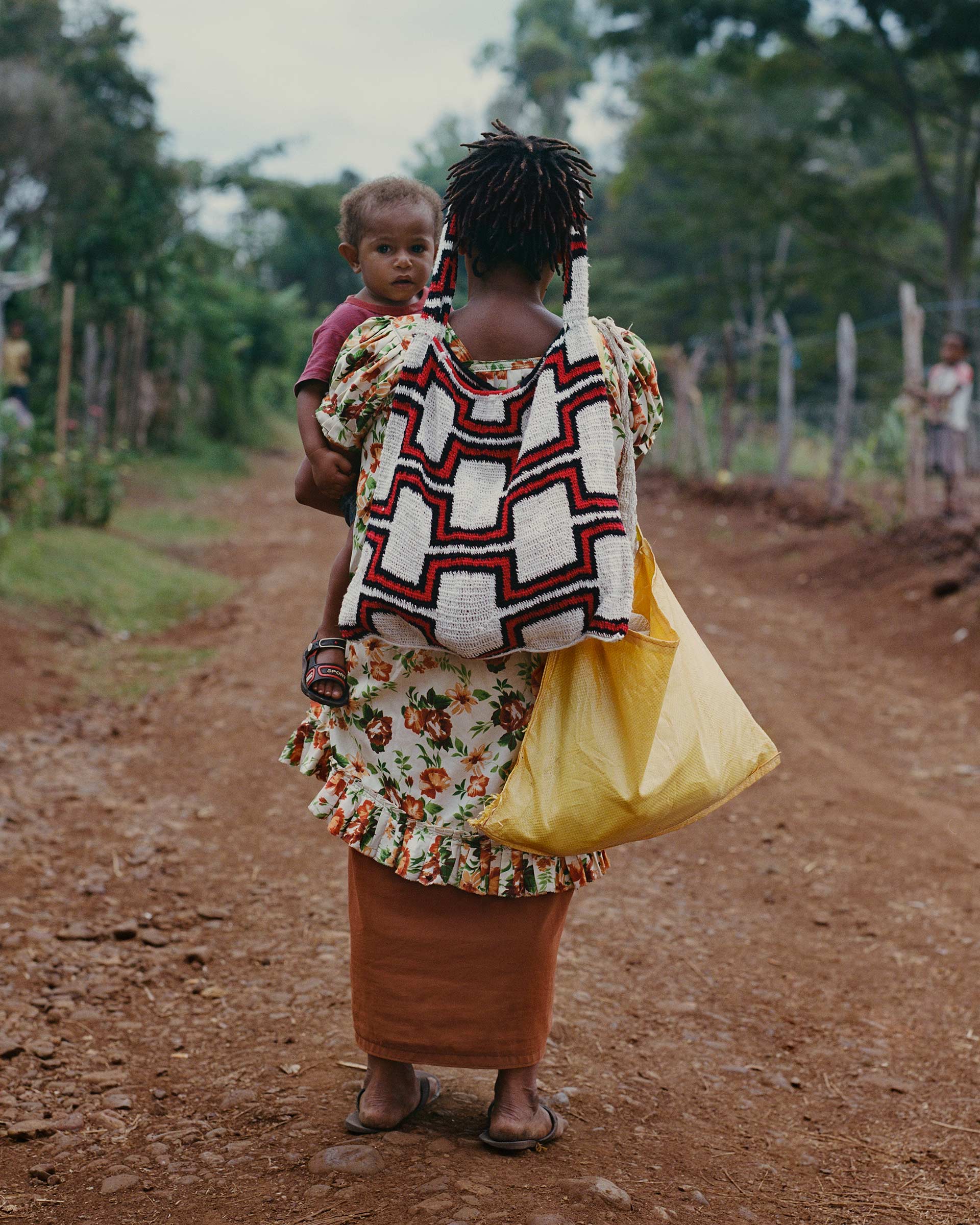 Mother carries child on back in rural setting. She wears a patterned bag and floral dress. Trees line a dirt road with distant figures.