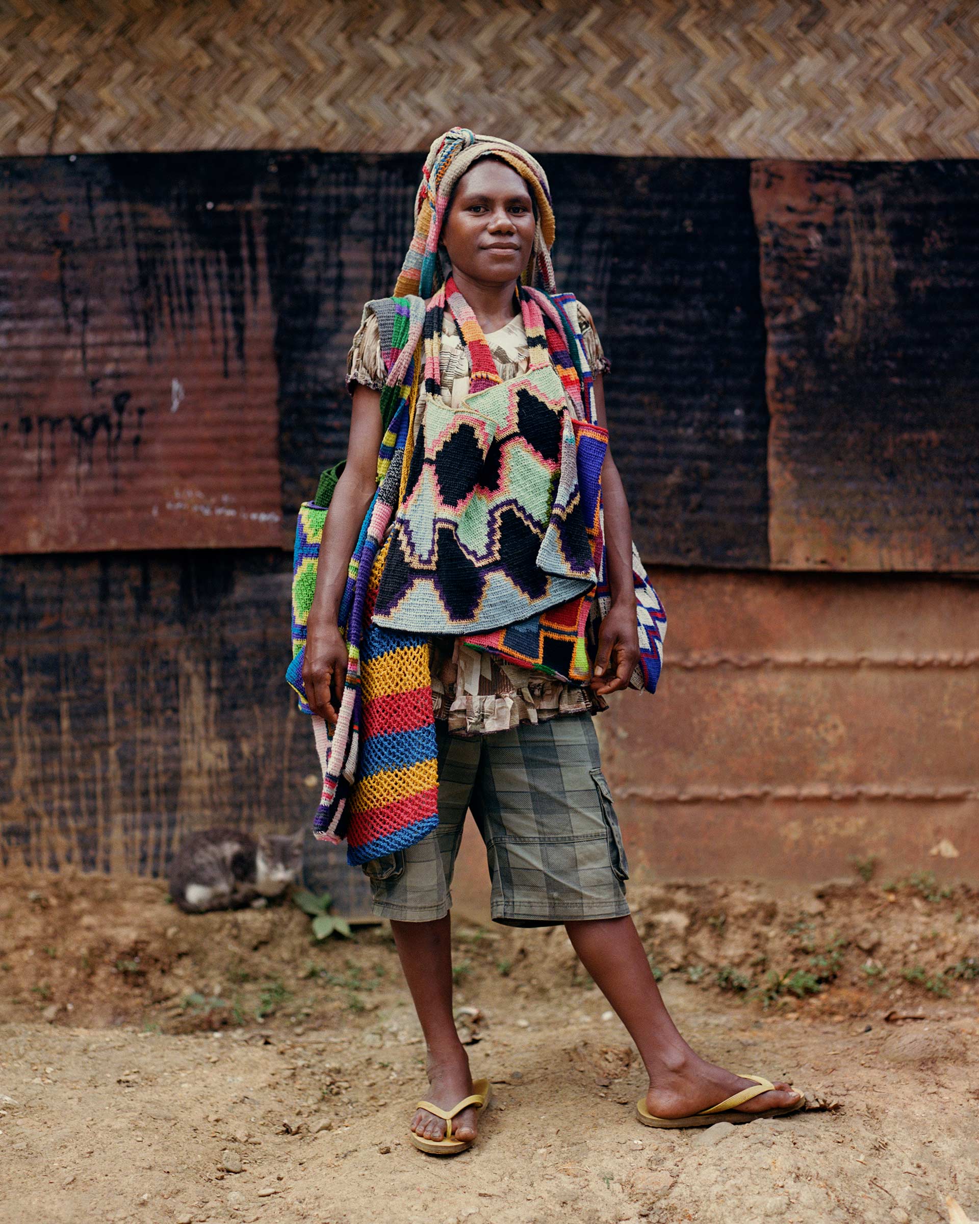 Full-length portrait of a woman wearing traditional clothing, including a headwrap and woven bags, standing in an outdoor setting.
