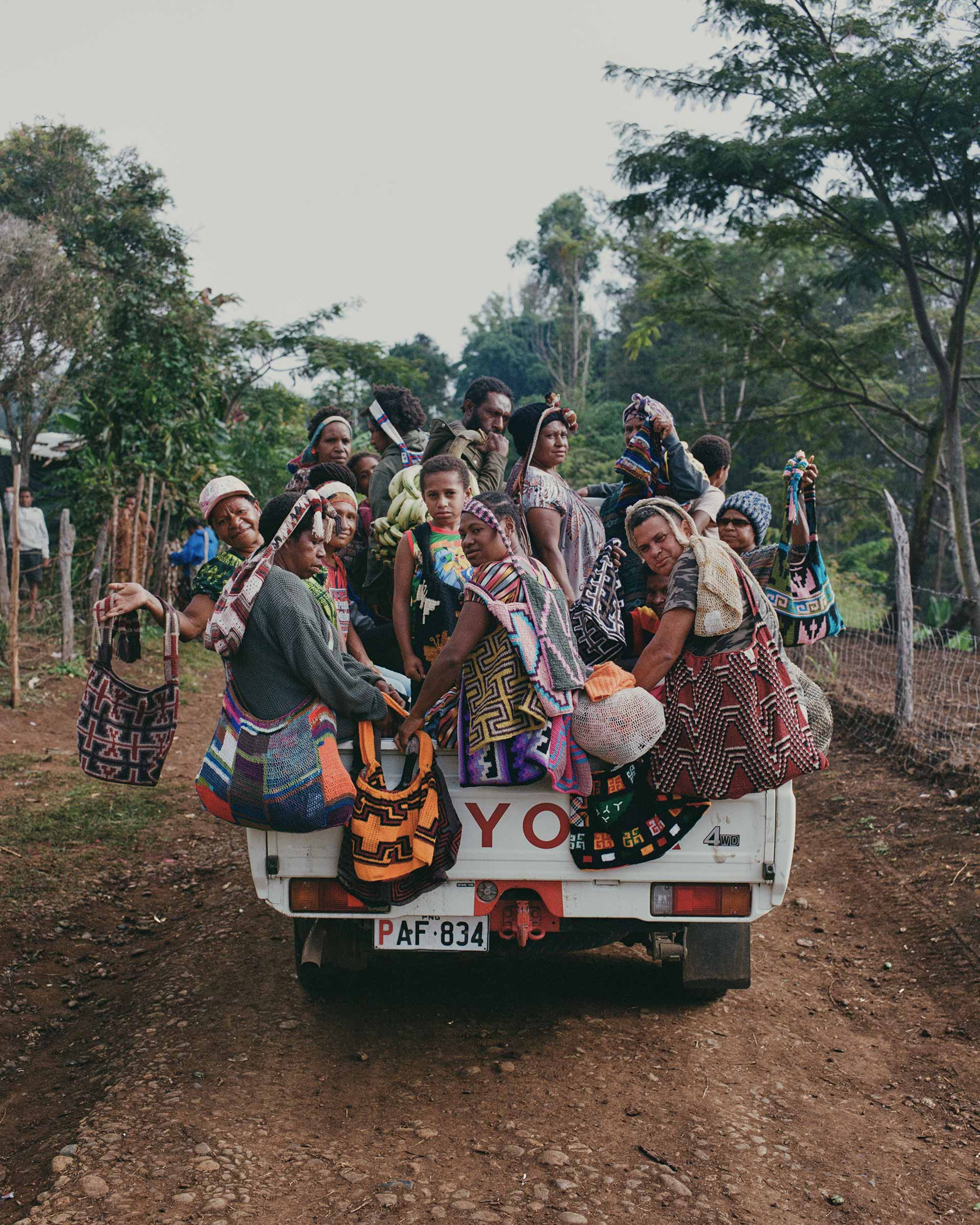 Group of indigenous women riding in the back of a pickup truck on a rural road, wearing colorful traditional clothes and carrying bags.