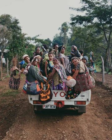 Group of indigenous women riding in the back of a pickup truck on a rural road, wearing colorful traditional clothes and carrying bags.