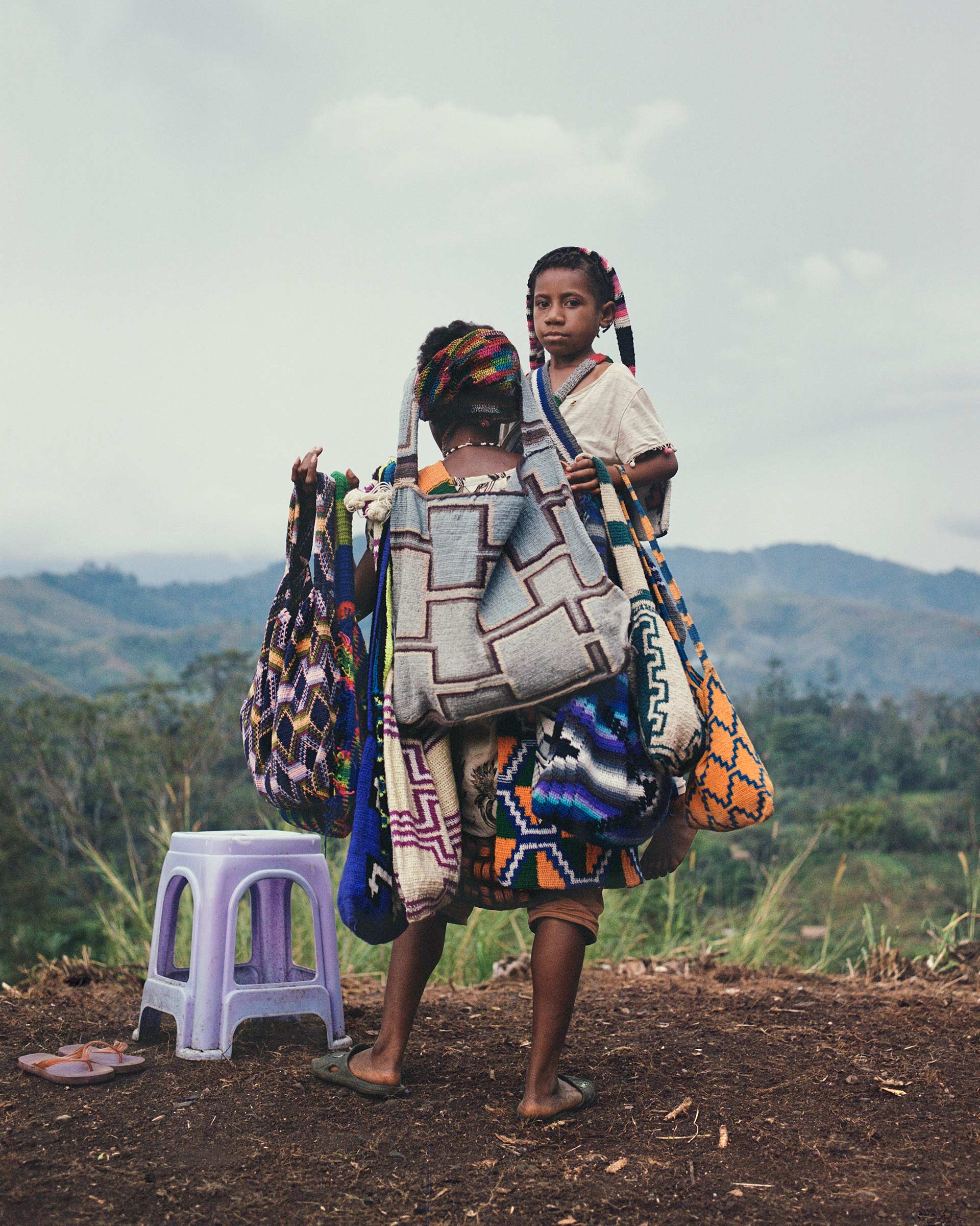 Two young girls are carrying colorful woven bags in a rural outdoor setting, with a stool and distant hills visible.
