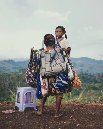 Two young girls are carrying colorful woven bags in a rural outdoor setting, with a stool and distant hills visible.