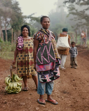 Two women standing on a dirt road, one holding a bunch of green bananas and the other wearing a patterned woven bag