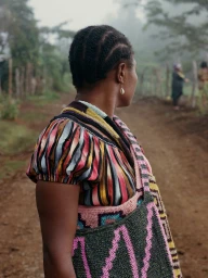 Profile of a woman with braided hair wearing a striped top and knitted patterned bag on a dirt path