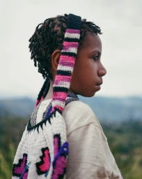Profile portrait of a young child with locs wearing a patterned crocheted bag strap across the shoulder against an outdoor backdrop.