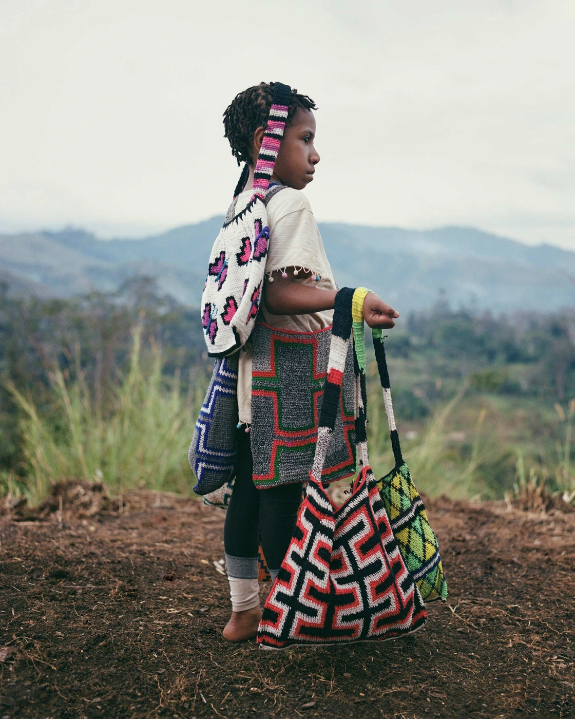 Child standing outdoors carrying multiple colourful patterned woven shoulder bags for Among Equals
