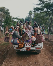 Group of people riding in the back of a white pickup truck on a dirt road, holding patterned woven bags.