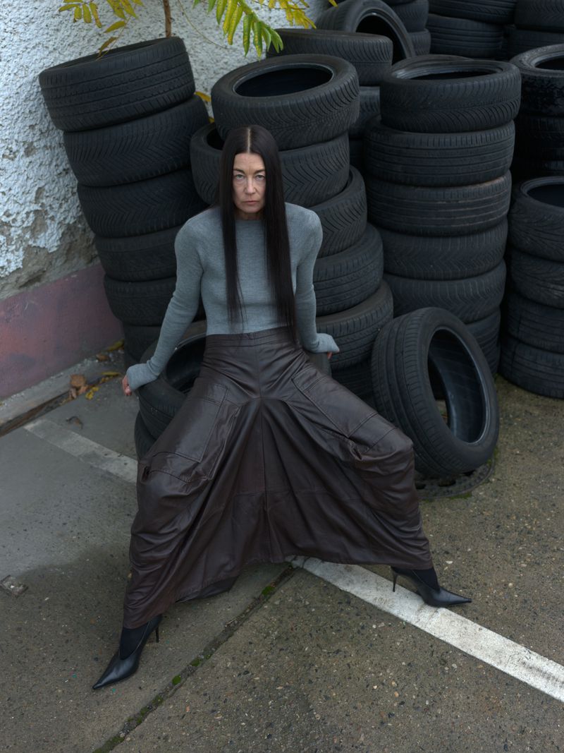 An adult woman sits on a stack of tires, wearing a gray top, brown harem pants, and black pointed-toe heels in an outdoor setting.