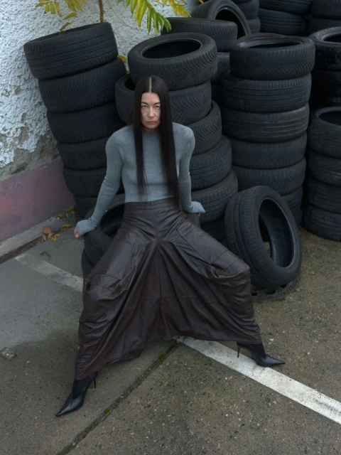 An adult woman sits on a stack of tires, wearing a gray top, brown harem pants, and black pointed-toe heels in an outdoor setting.