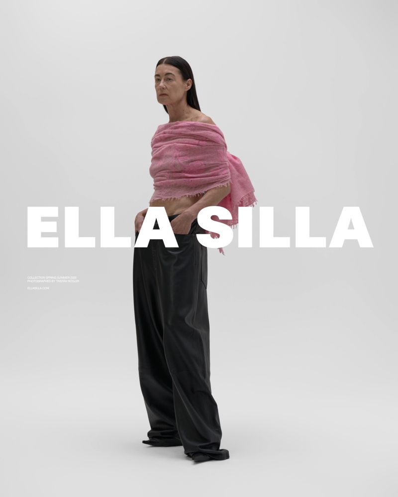 Fashion shot of an adult woman in baggy black pants and pink shawl, standing indoors against a white backdrop, with the text ELLA SILLA.