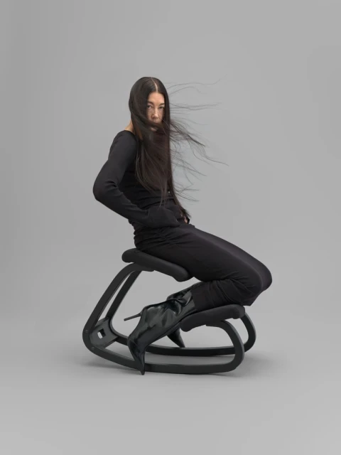 Adult woman with long hair sits on a unique ergonomic chair in a studio, posing in a black, long-sleeve jumpsuit and black heeled boots.