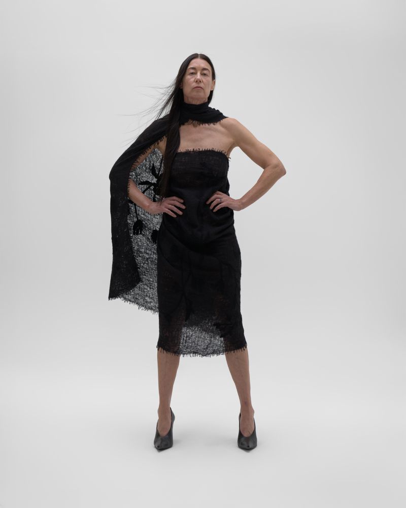 Woman in black lace dress, cape, and heels poses in a photo studio against a neutral background, hands on hips, looking at the camera.