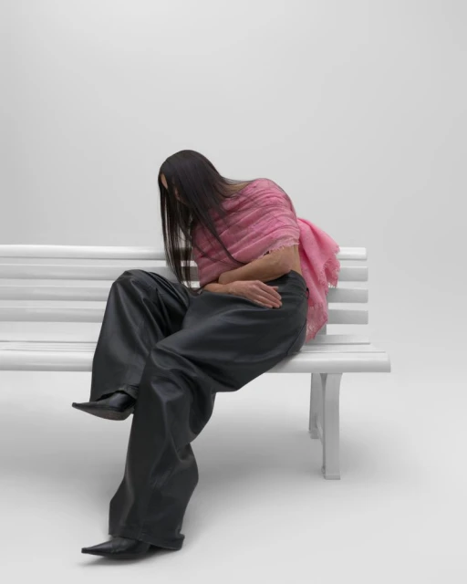 A person sitting on a white bench, wearing black pants and a pink scarf, with long dark hair against a plain white background.
