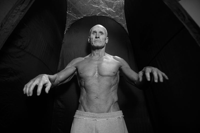 Older man with muscular physique poses shirtless against a dark background with arms outstretched in a dramatic black and white portrait.