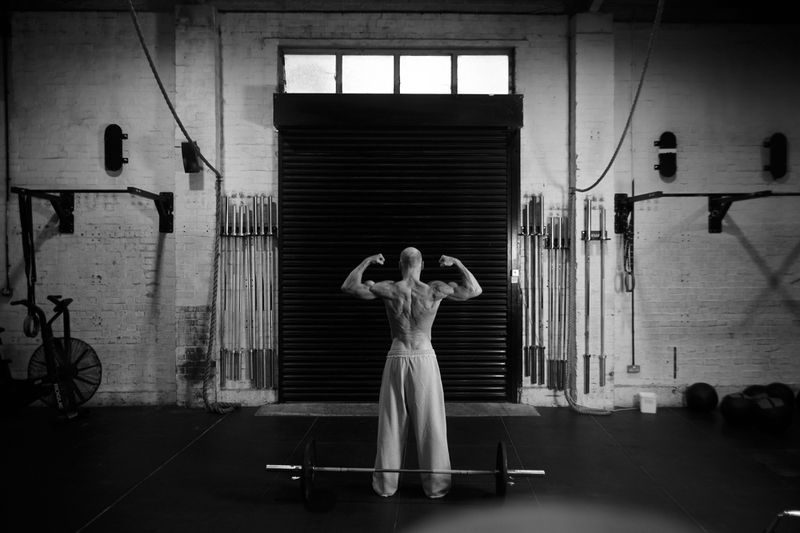 Muscular man flexing back muscles in an indoor gym with weights and fitness equipment against a closed black shutter door.
