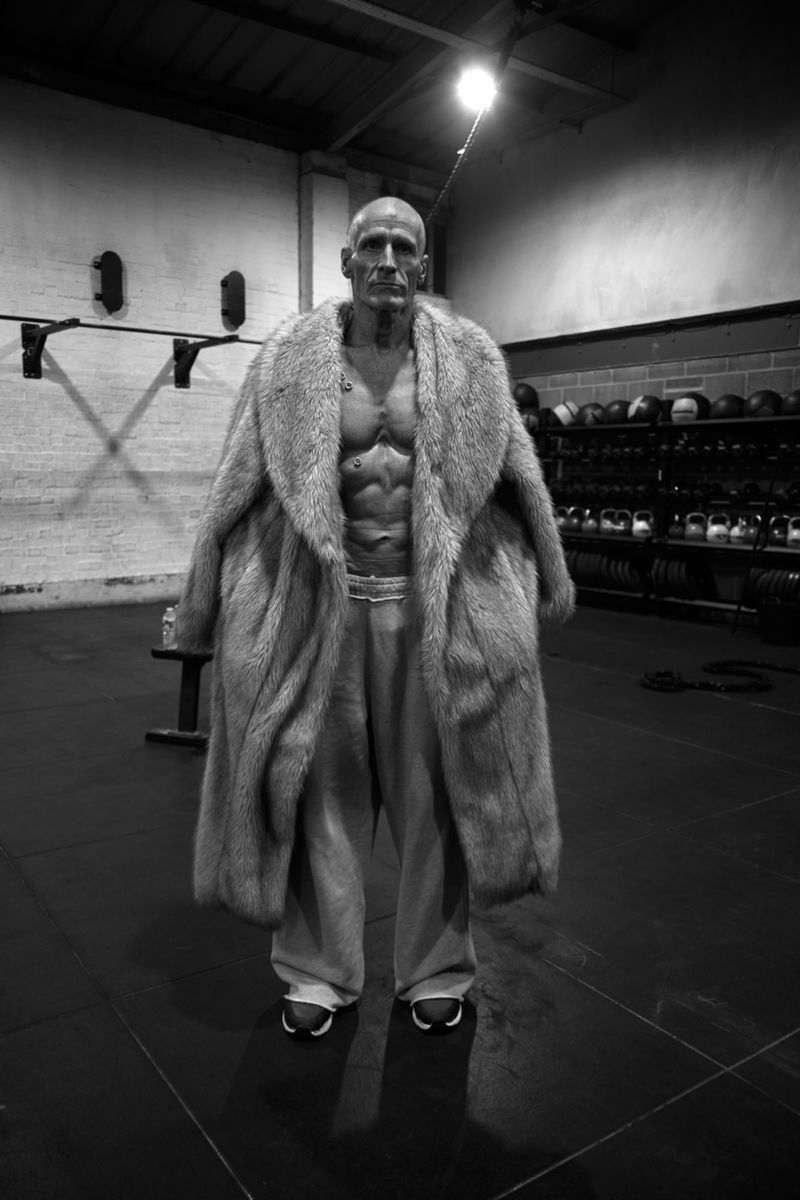 Adult male with muscular physique poses in an indoor gym setting wearing a fur coat, pants, and sneakers in a full-length shot.