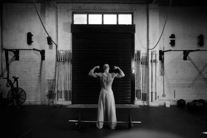 Muscular man flexing his back muscles in a gym setting with a barbell on the floor.