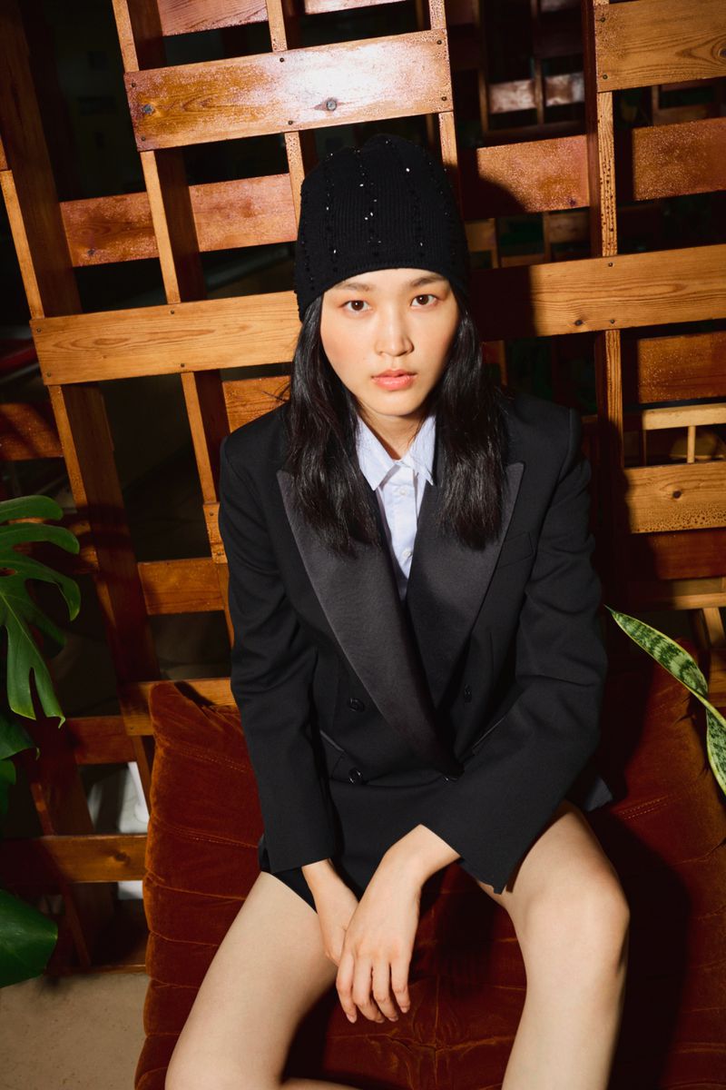 Model wearing a black blazer and beanie seated against a wooden structure for MAX&Co.