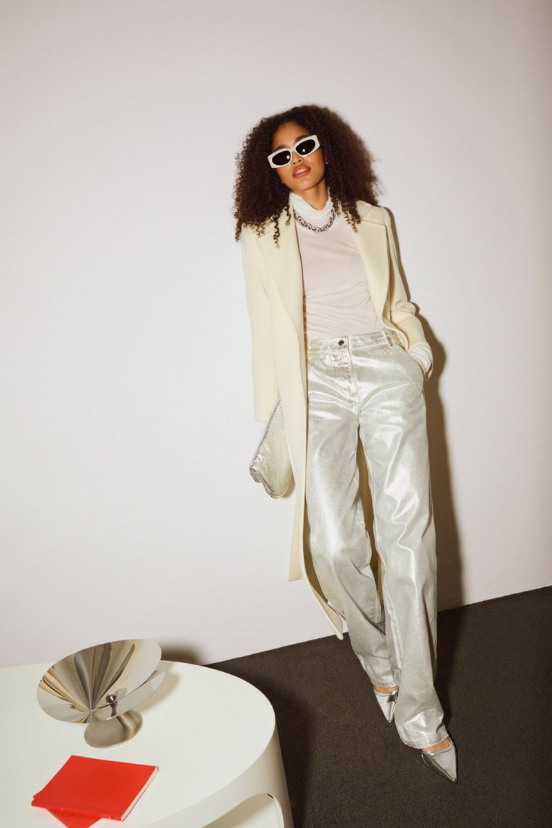 A woman with curly hair poses in a cream coat and silver pants, accessorized with white sunglasses and jewelry, against a light background.