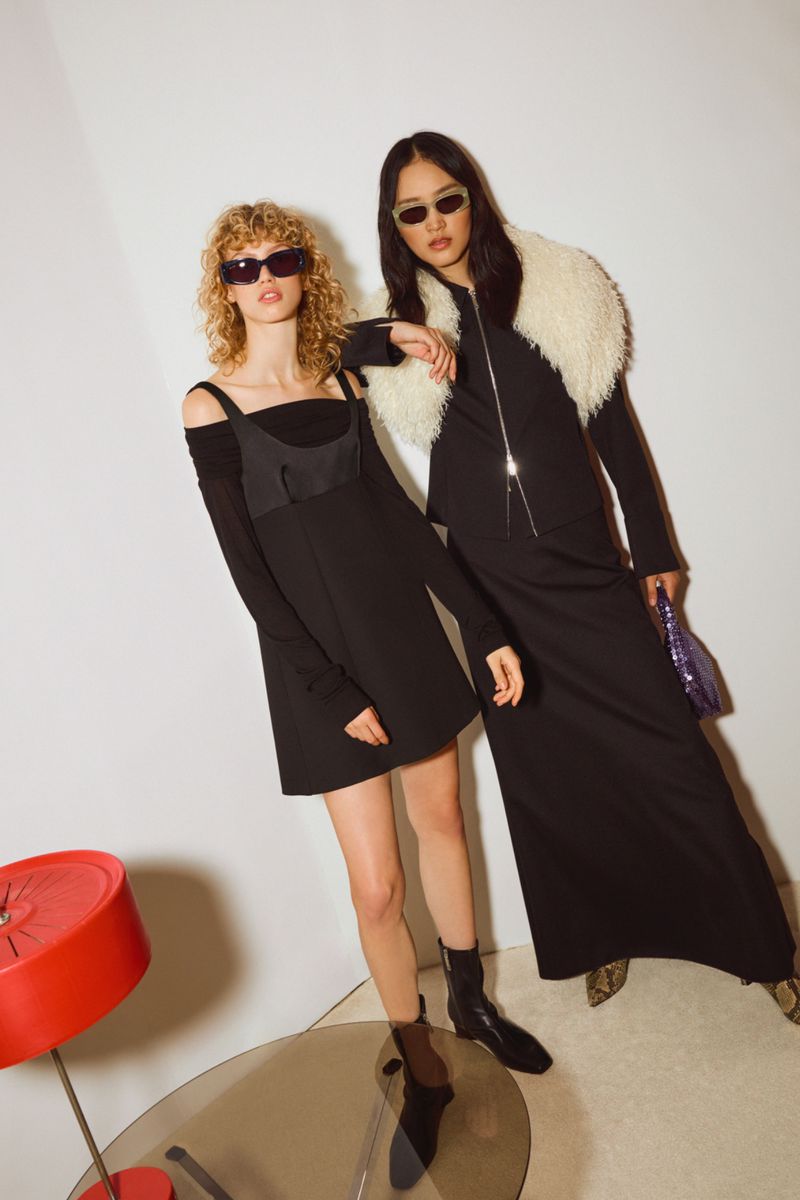 Two women pose indoors wearing black dresses and sunglasses; one in a long dress with a white fur collar. Modern fashion photography.