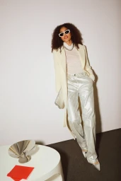 Model wearing white sunglasses, cream coat, and metallic pants leaning against a neutral studio wall.