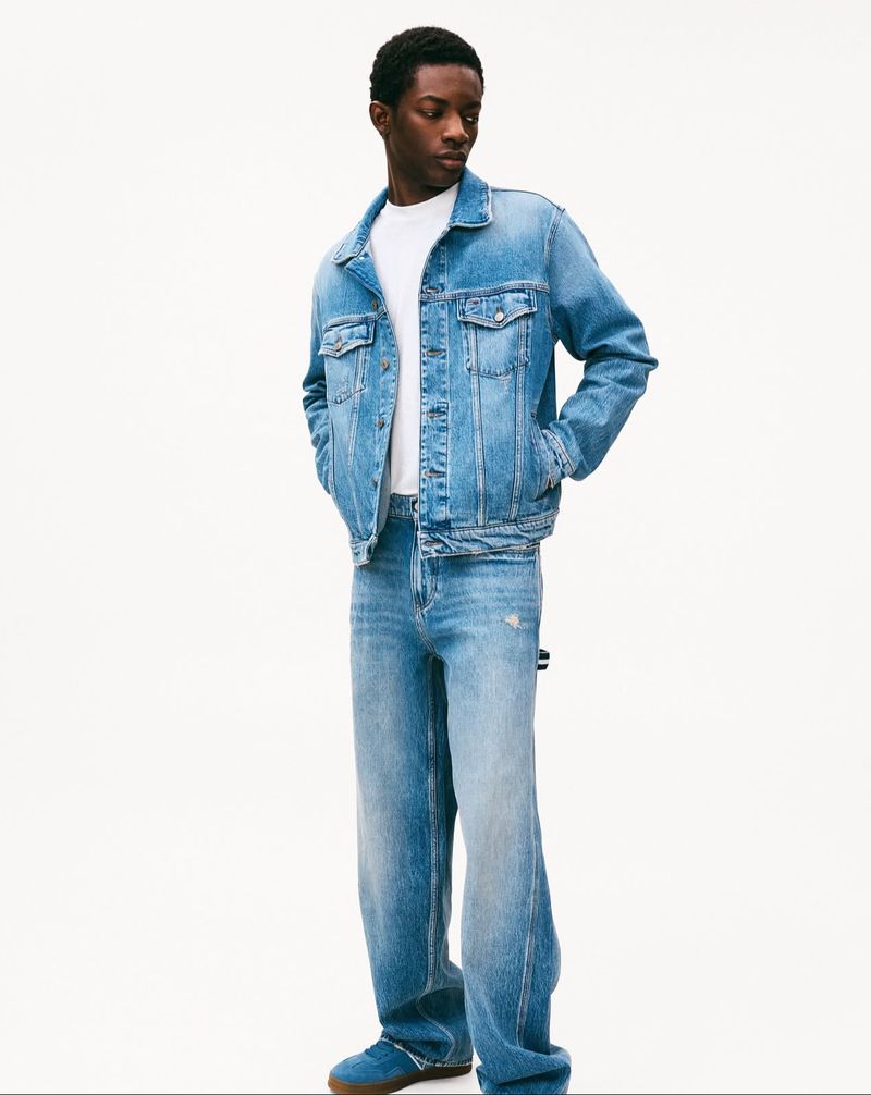 Full-length shot of a Black male model in a denim jacket, white tee, loose jeans, and blue shoes, posing against a white background in a studio.