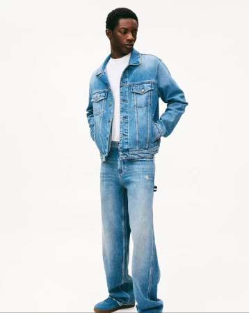 Full-length shot of a Black male model in a denim jacket, white tee, loose jeans, and blue shoes, posing against a white background in a studio.