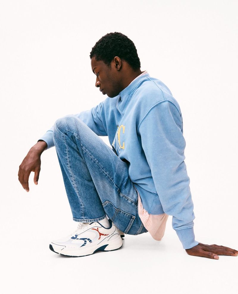 Man in light blue sweatshirt and jeans squats indoors on a white surface, wearing athletic shoes, looking downward in a fashion shot.