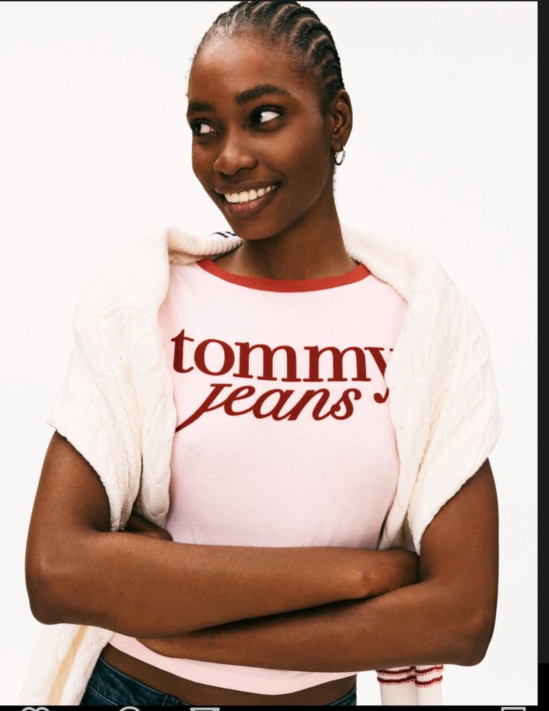Portrait of a smiling African-American woman wearing a pink Tommy Jeans T-shirt and a white sweater with arms crossed, set against a white background.
