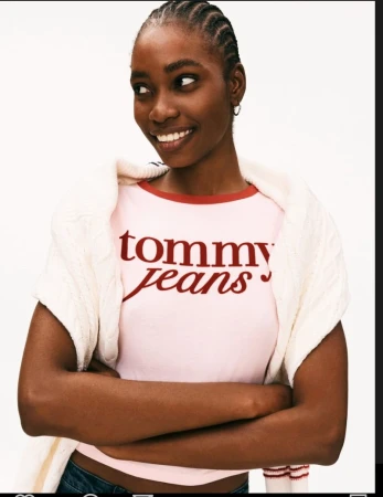 Portrait of a smiling African-American woman wearing a pink Tommy Jeans T-shirt and a white sweater with arms crossed, set against a white background.