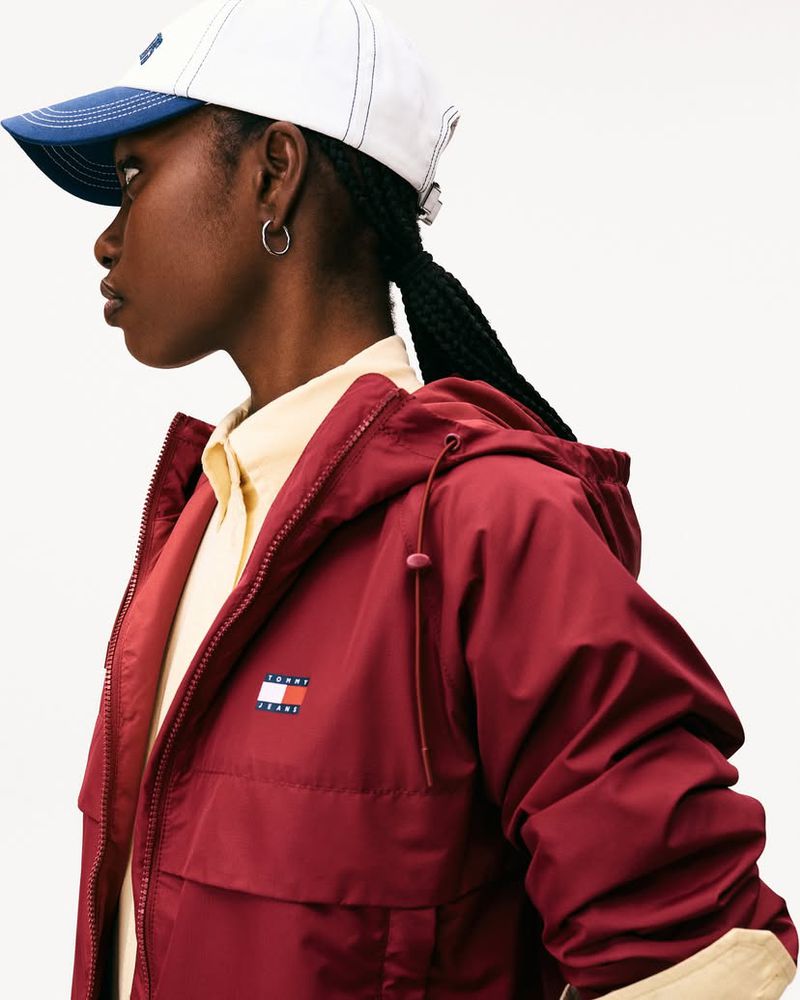 A young adult woman in a stylish red jacket and white baseball cap, photographed in a studio setting with a clean, neutral background.