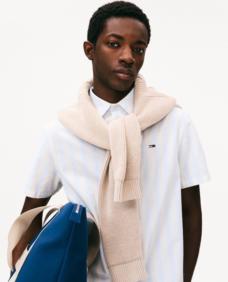 A young Black man is posing indoors, wearing a white shirt with a beige sweater draped over his shoulders and a blue bag nearby.
