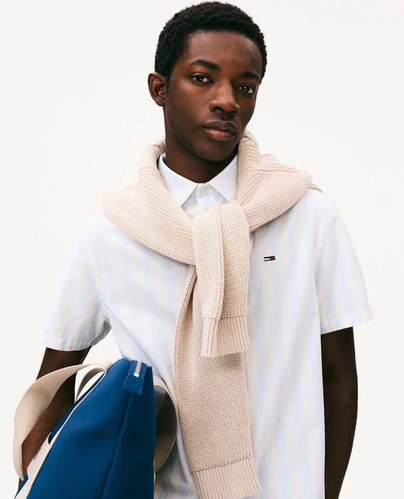 Black man holding blue bag, wearing white shirt and cream sweater tied over shoulders