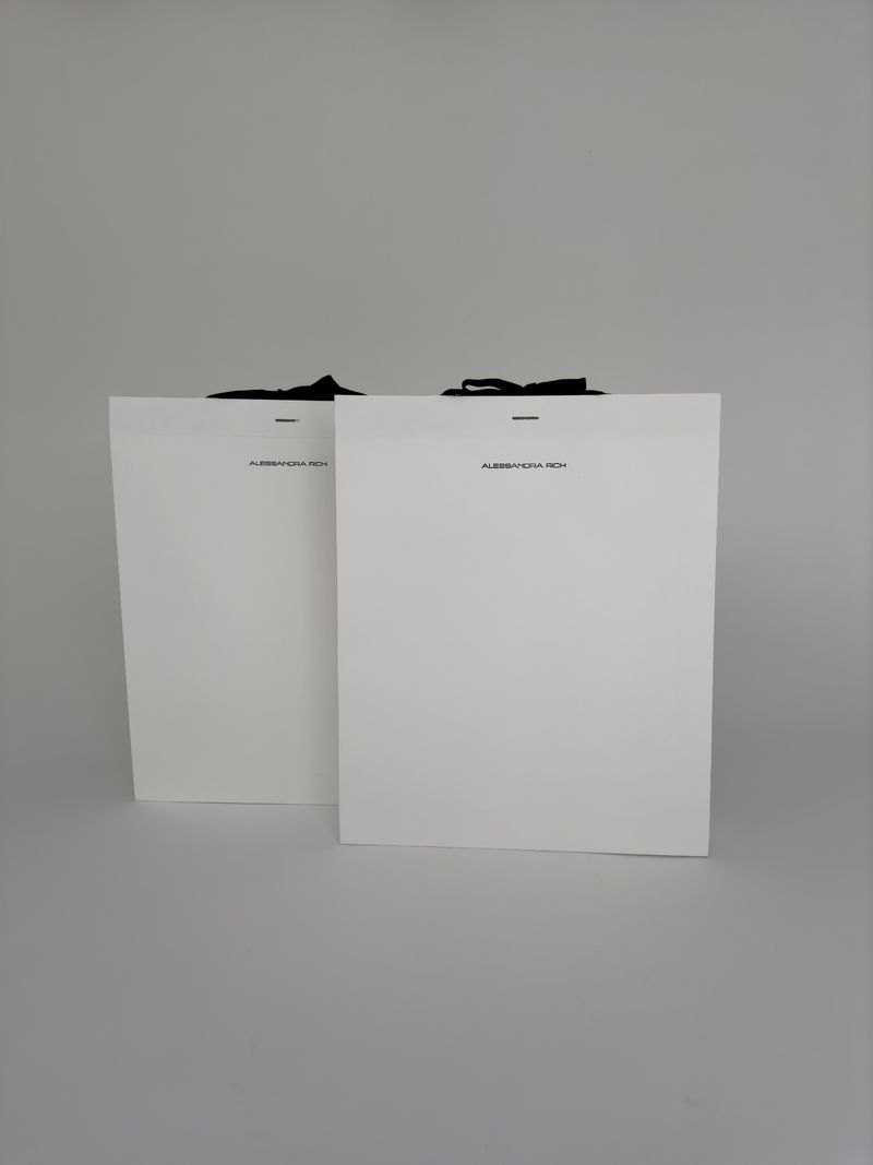 Two white shopping bags stand side-by-side, with black ribbon handles, against a solid light gray background in a studio setting.