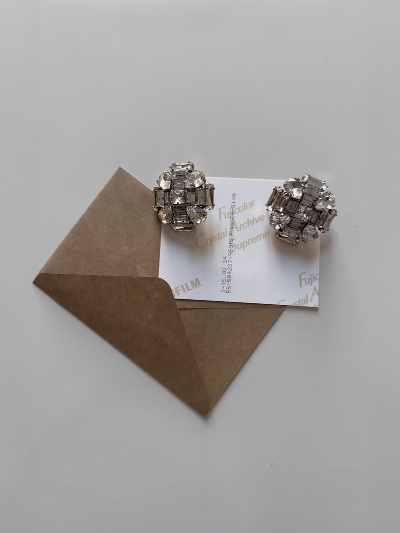Pair of rhinestone-encrusted silver earrings displayed with a brown envelope and film paper on a white surface in a close-up shot.