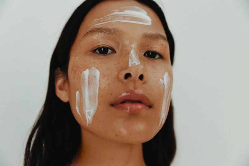 Close-up of a tan-skinned young woman with white cream on her face, looking directly at the camera against a neutral background.