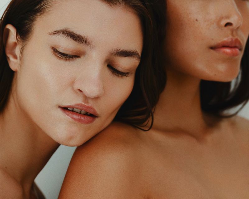 Close-up studio portrait of two women with different skin tones, one with closed eyes resting on the other's shoulder.