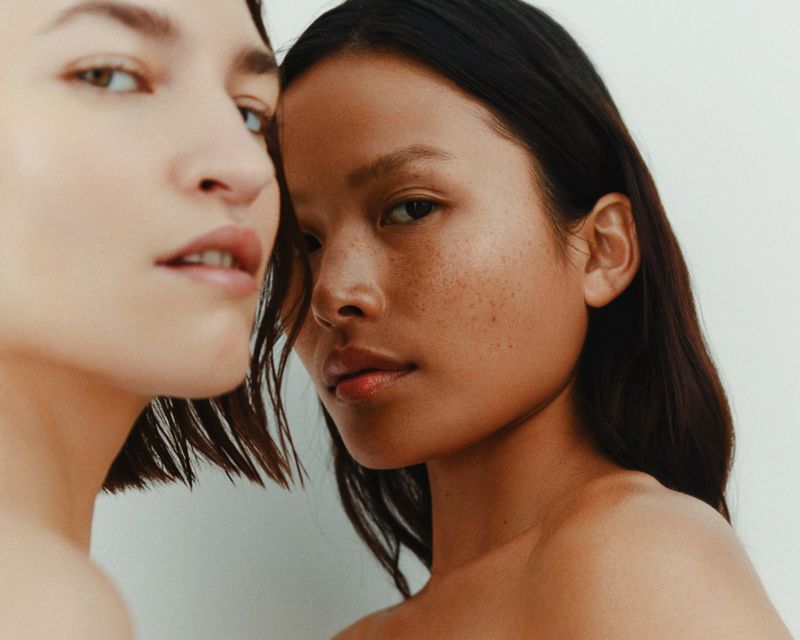 Close-up portrait of two young adult females with fair and medium skin tones, dark hair, and bare shoulders against a plain white background.