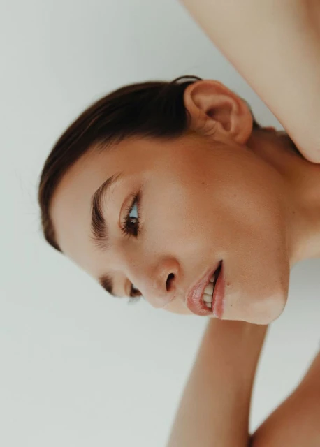 Headshot of a woman with dark hair, fair skin, light eyes, arms behind her head, and white backdrop in soft, natural light.