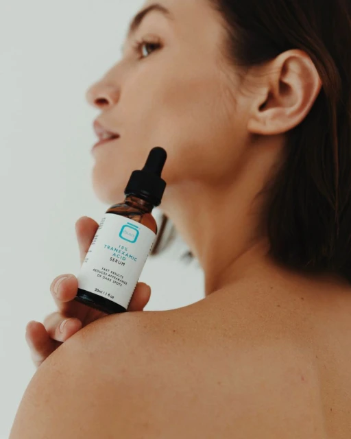 Woman's skincare routine: Adult woman holds a serum bottle in front of her shoulder in a close-up studio shot on white background.
