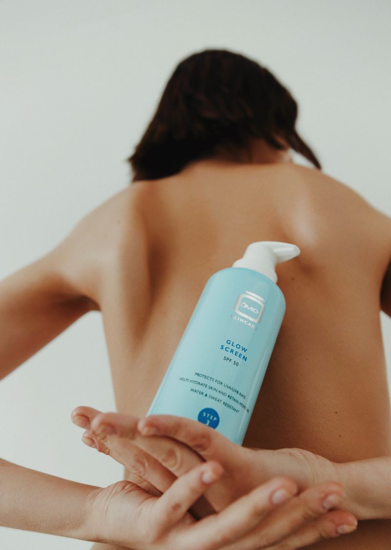Woman holding a light blue bottle of 'Glow Screen' sunscreen product in front of her bare back in a minimalist studio setting.
