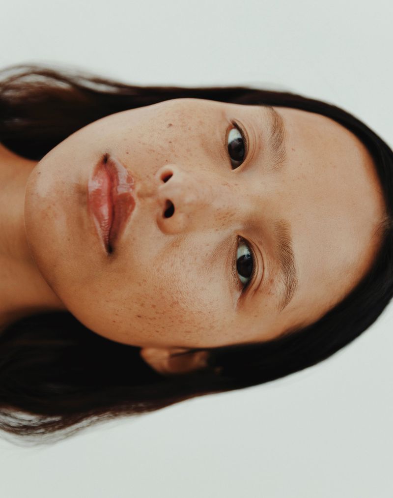Close-up beauty portrait of a model with dark hair and freckles against a neutral background for OMIC Beauty Spring 2025.