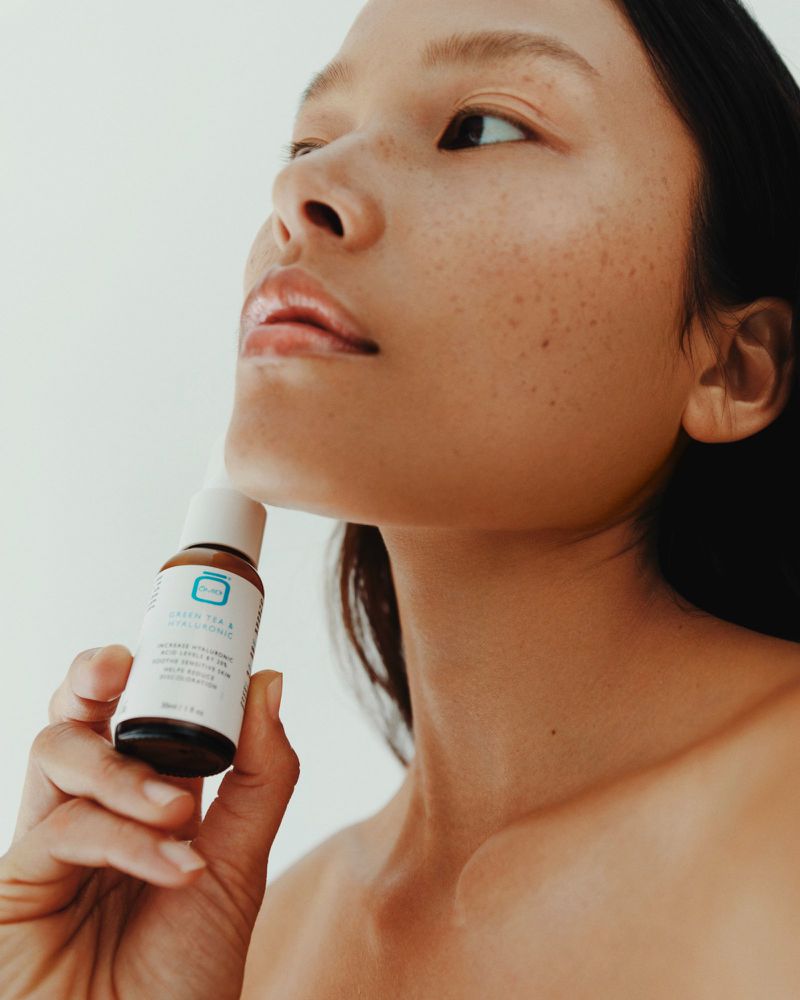 Asian woman applies a skincare product. The serum bottle is in her hand, with a light background and focus on beauty treatment.