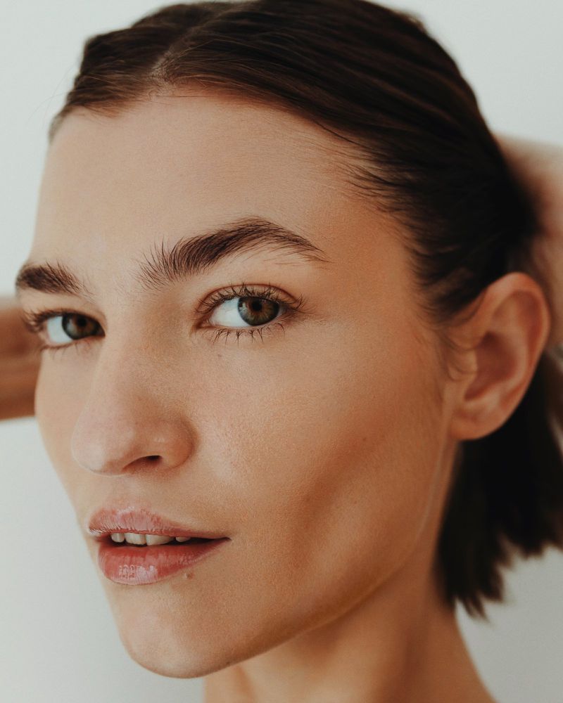 Close-up of a young adult woman with tan skin and brown hair, looking at the camera against a plain white background.