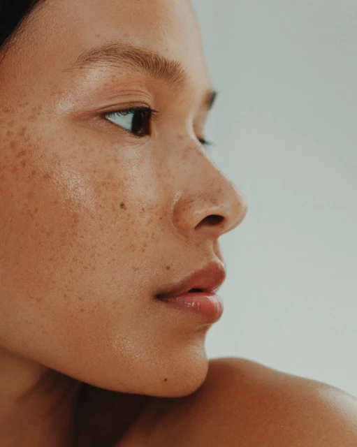 Close-up portrait of a young adult female with tanned skin and freckles, looking off-camera against a soft, neutral background.