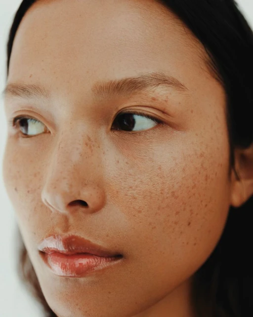 Close-up portrait of a woman with freckles and glossy lips looking off to the side against a neutral background, beauty concept.